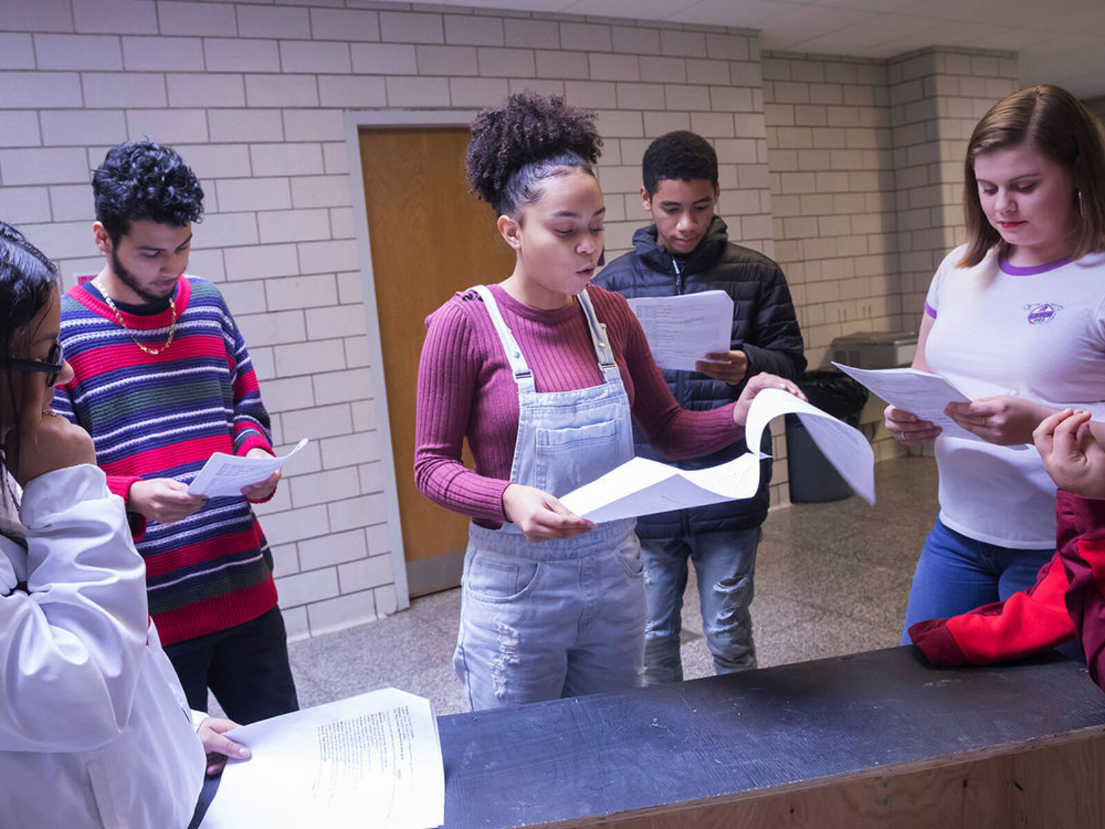 Six Mount Holyoke High School students stand together in a hallway. Facing the camera, they read from paper scripts during a small group rehearsal.