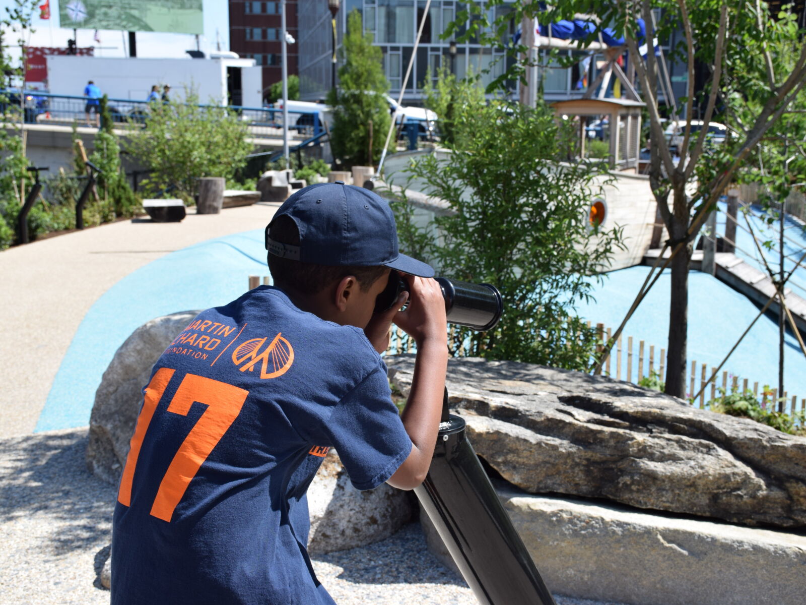 A child looks through a periscope installed at Martin's Park along the Boston Waterfront. He wears a navy blue shirt and baseball cap. He is surrounded by green trees and play structures.