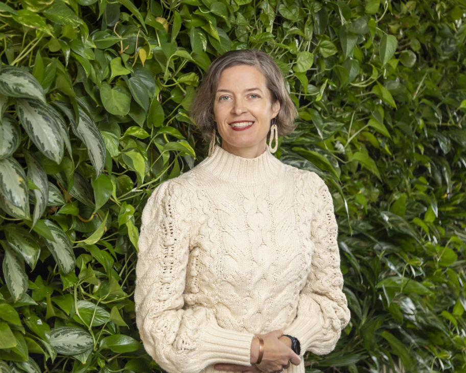 White woman with short brown hair wearing a thick, cream sweater smiles and stands in front of a green plant wall