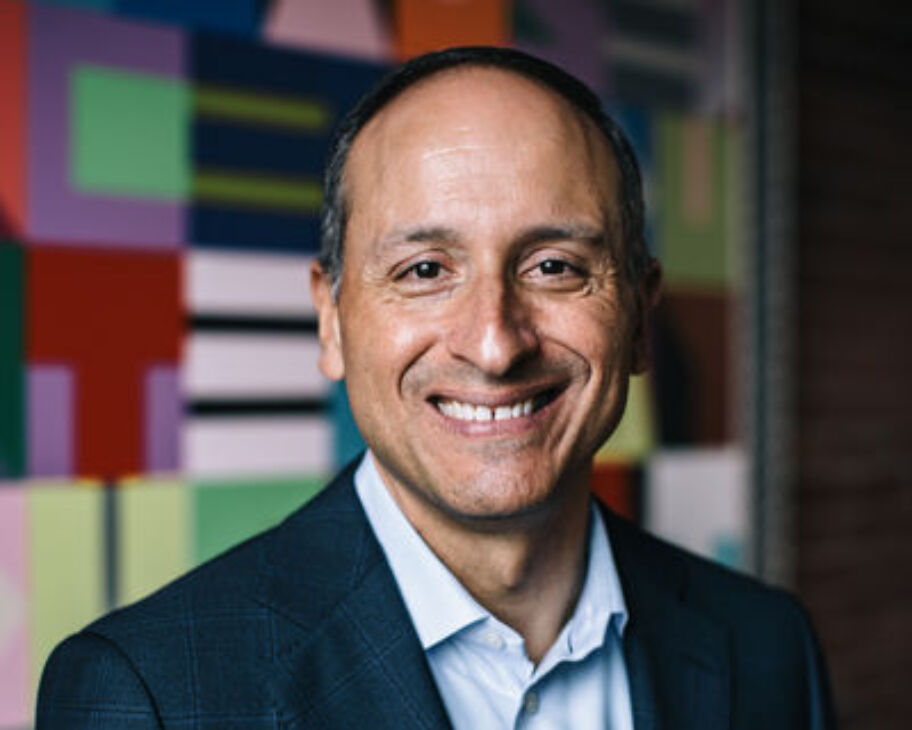 Jim Canales, Barr Foundation CEO stands in front of an artwork by Jeffrey Gibson that shows a patchwork of colorful lines and shapes. He smiles broadly and wears a blue suit jacket and shirt.