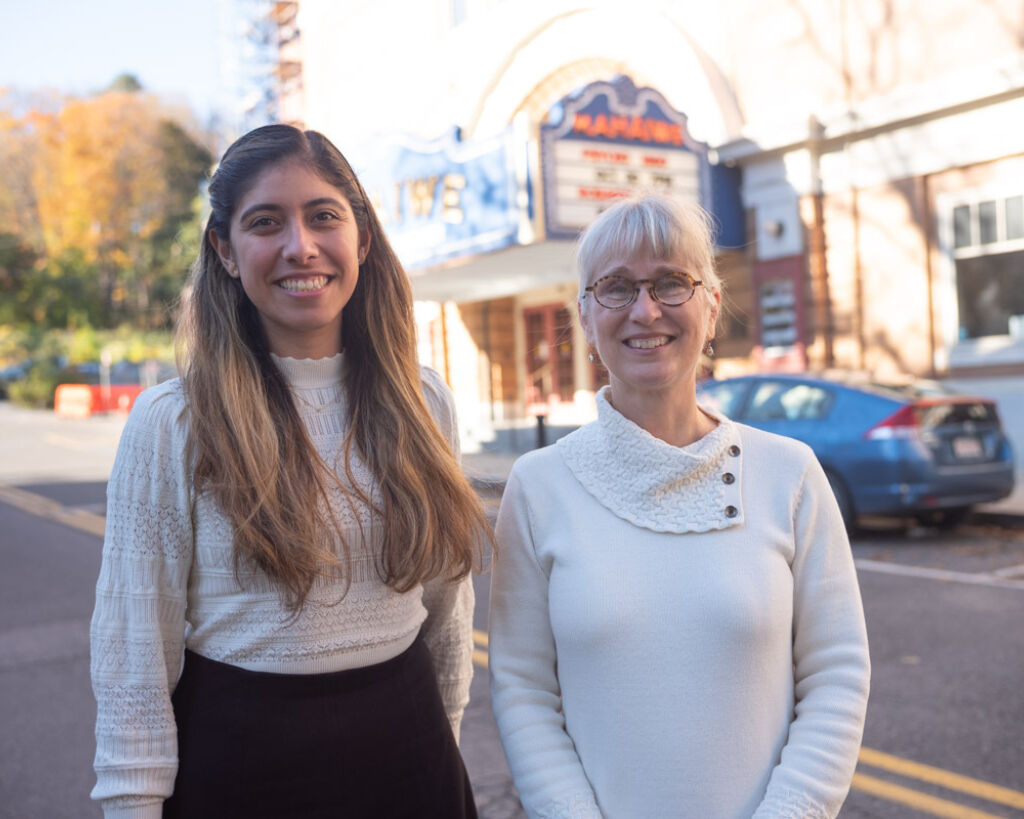 Two smiling women stand outside, across the street from the Mahaiwe Performing Arts Center. One older white woman is the executive director of the center and one younger latina woman is a member of the center's Spanish language advisory newtork.