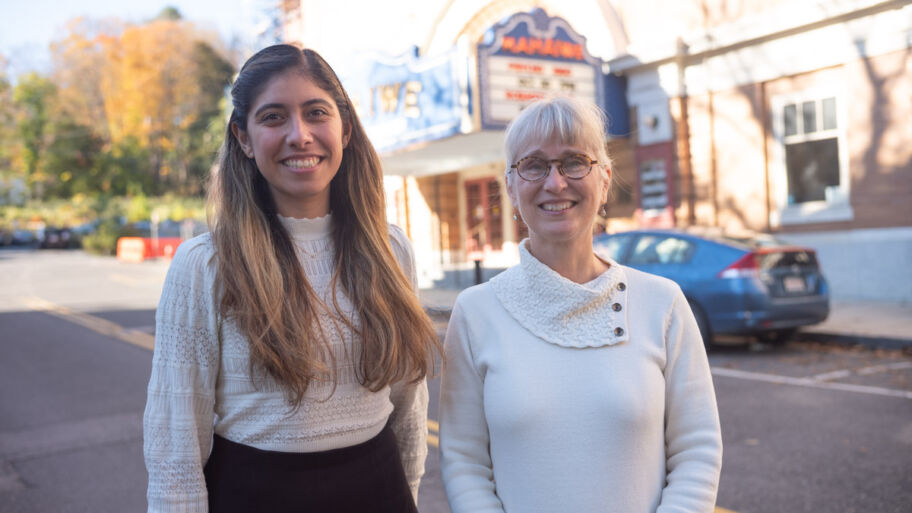 Two smiling women stand outside, across the street from the Mahaiwe Performing Arts Center. One older white woman is the executive director of the center and one younger latina woman is a member of the center's Spanish language advisory newtork.