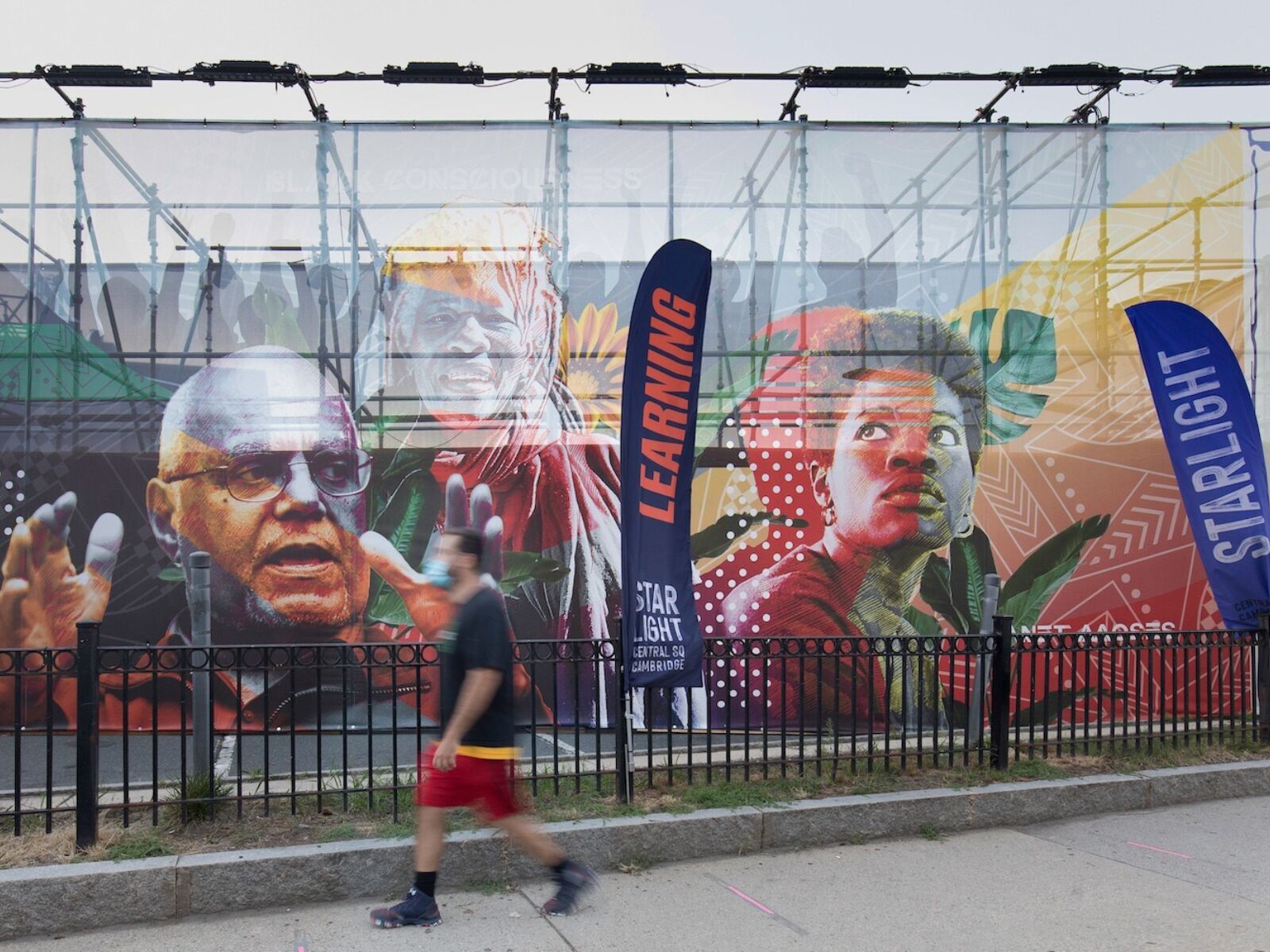 In Cambridge, Massachusetts, a person walks along a sidewalk, blurred in motion. Behind him, a fenced in parking lot has been covered with large-scale graphic art of Black Bostonians. Large standing flags along the route read "Learning" and "Starlight."