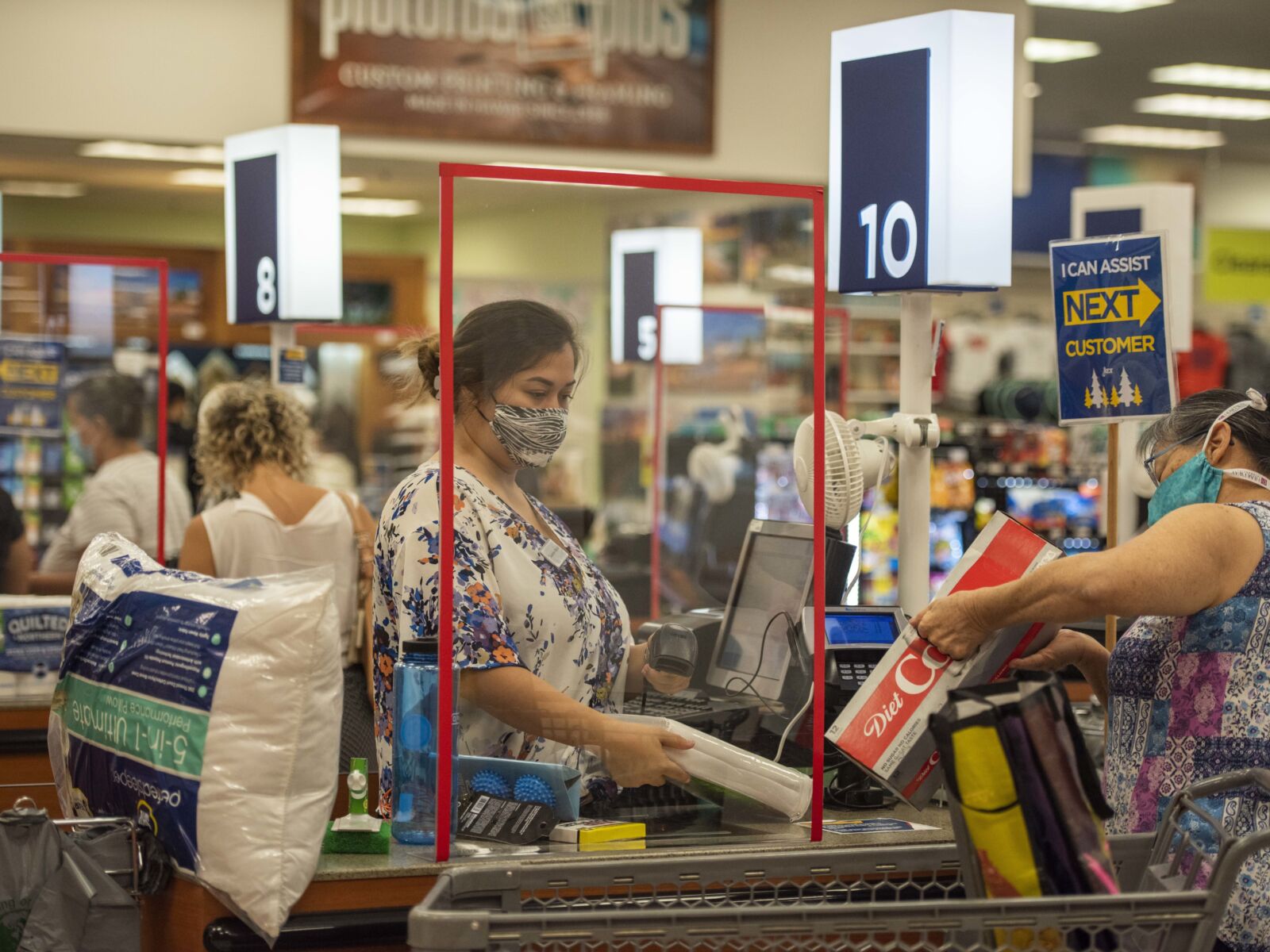 A scene from a busy check-out area in a grocery store during the height of the COVID-19 pandemic. Patrons wear mostly homemade masks. Cashiers stand behind plastic shields as they scan items for customers.