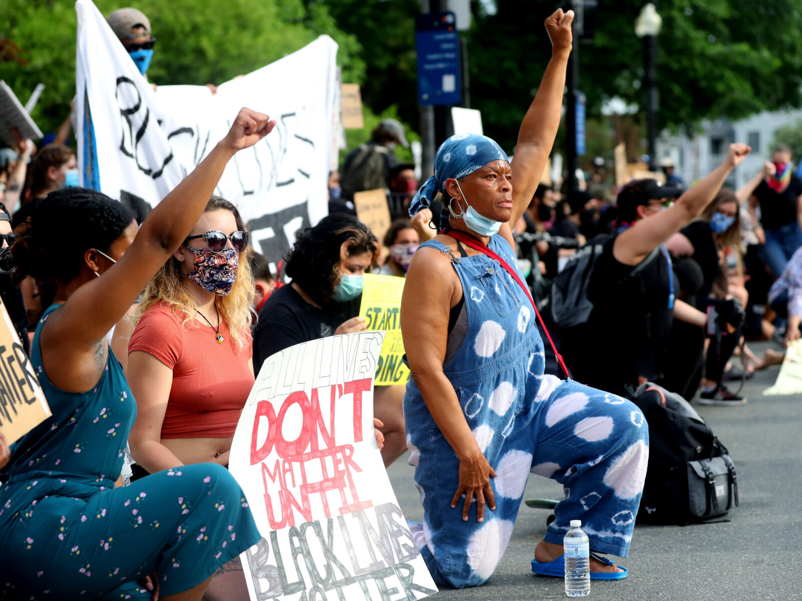 Protesters kneel during a Black Lives Matter rally in Boston, Massachusetts. Many are masked and raise their fists or hold protest signs. A woman in the foreground is especially striking--she wears denim overalls and looks resolutely in front of her.