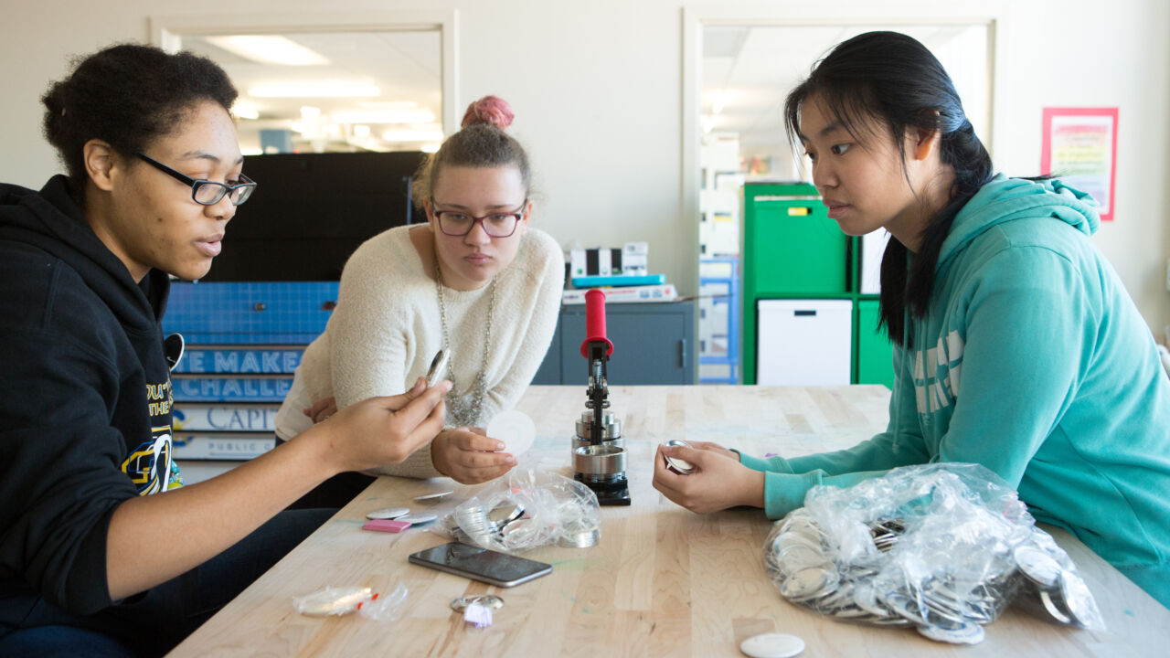 Three girls makes buttons in a high school classroom.
