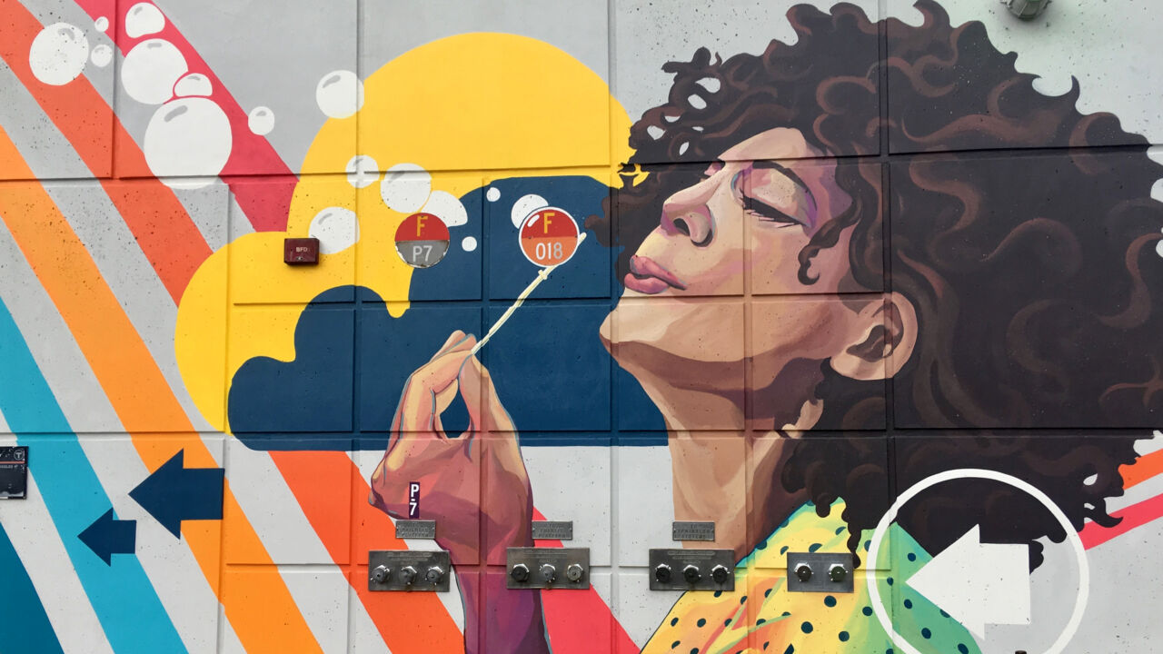 Colorful mural of woman with brown hair and red lipstick blowing white bubbles across a rainbow.