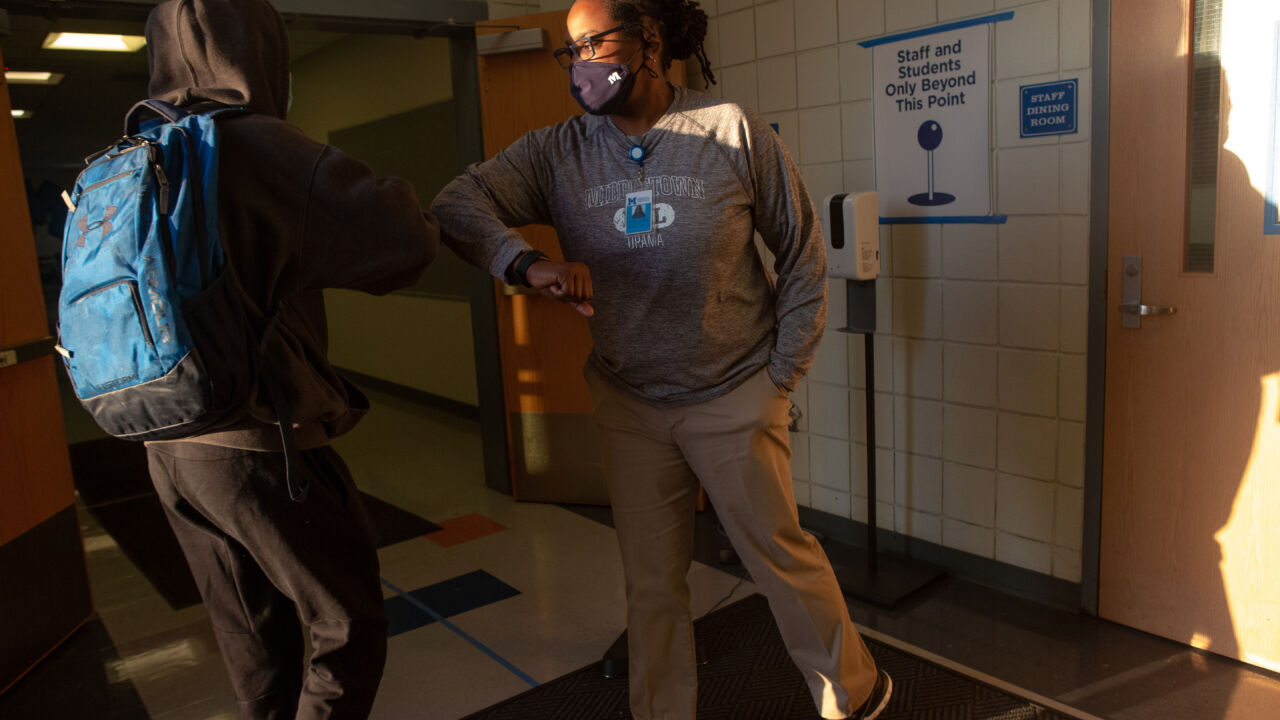 A masked teacher greets students coming into school.