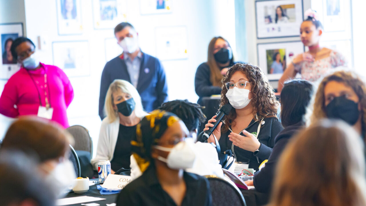 A masked speaker speaking to a room full of people at a spring convening.