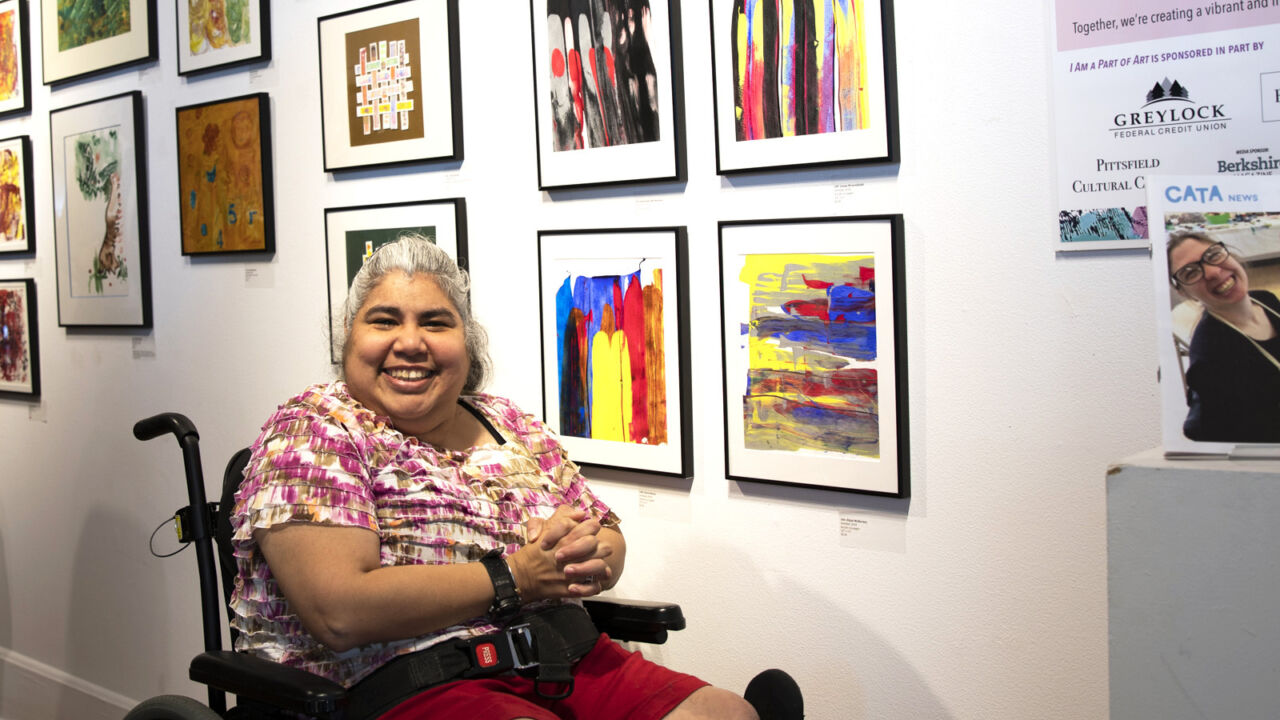 Woman sitting in a wheelchair in front of a wall of artwork, smiling