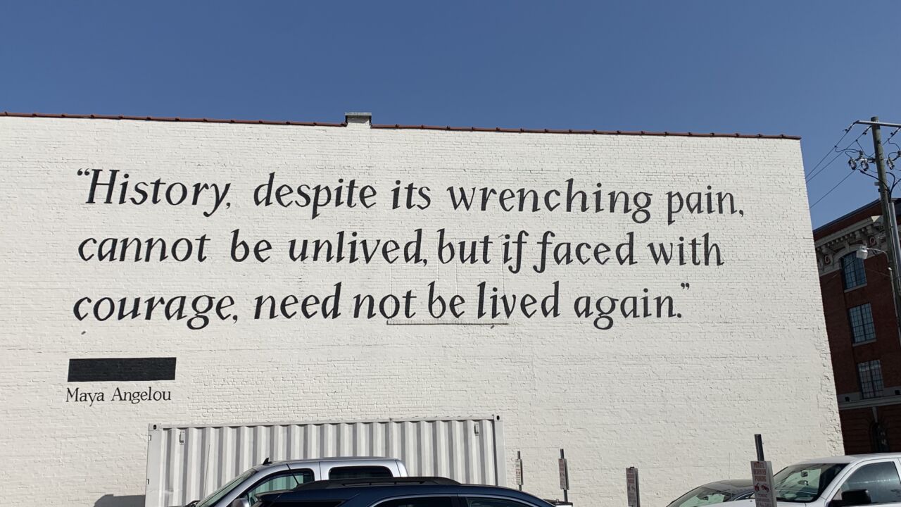 Black text on a white brick wall at the edge of a parking lot. It reads “History, despite its wrenching pain, cannot be unlived, but if faced with courage, need not be lived again.” over the name Maya Angelou
