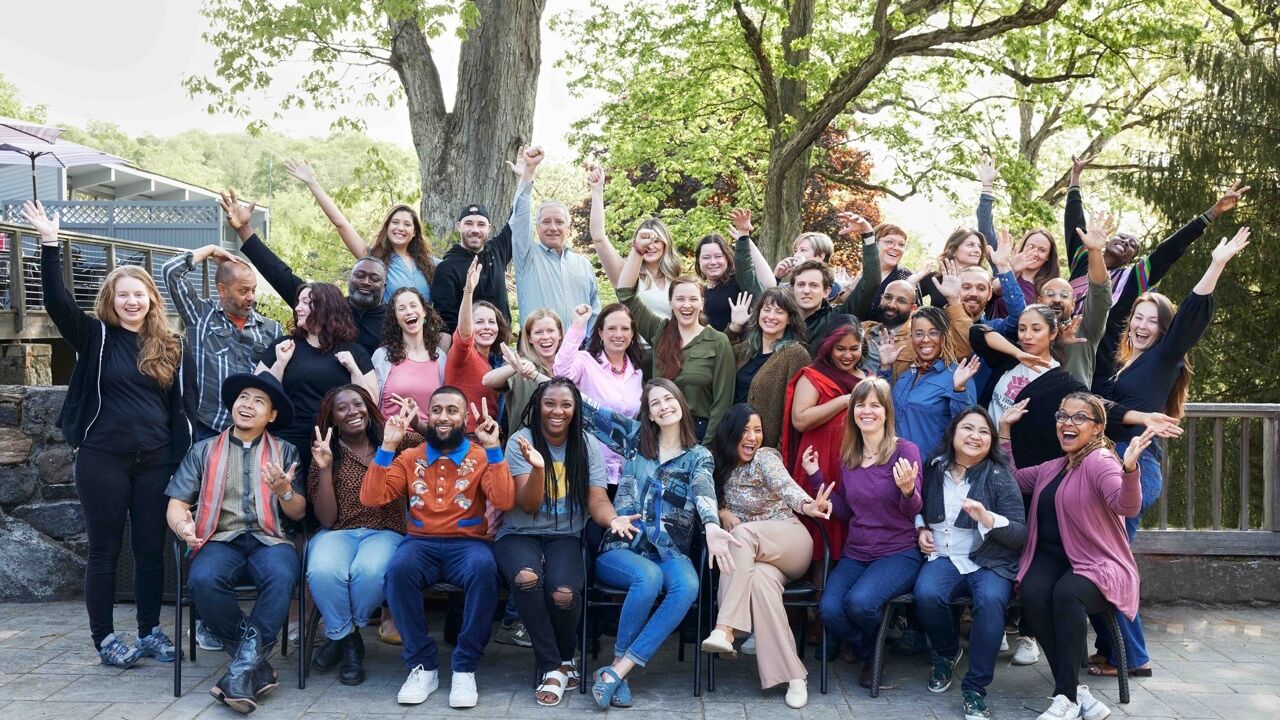 A diverse group of forty smiling people, many with hands raised, on a stone patio in front of green trees