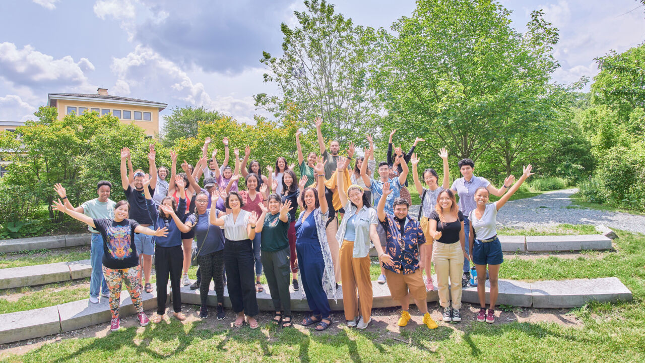 A group of people of color standing outside on green grass, acting excited with their arms in the air.