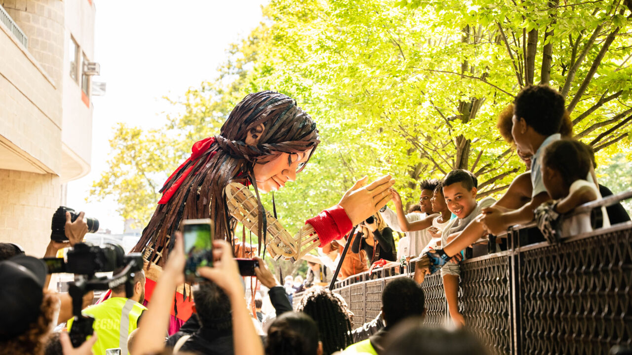 A large puppet of a 10-year-old Syrian girl named Little Amal interacts with a crowd.