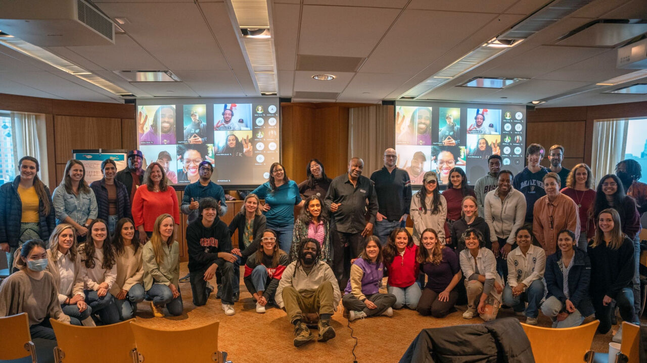 A diverse group of approximately 30 people smiling and posing in a conference room, standing in front of two projection screens with several additional people joining virtually.