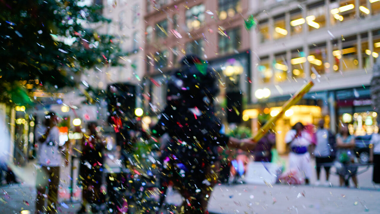 A blurry scene of performers in a public urban space with colorful confetti.