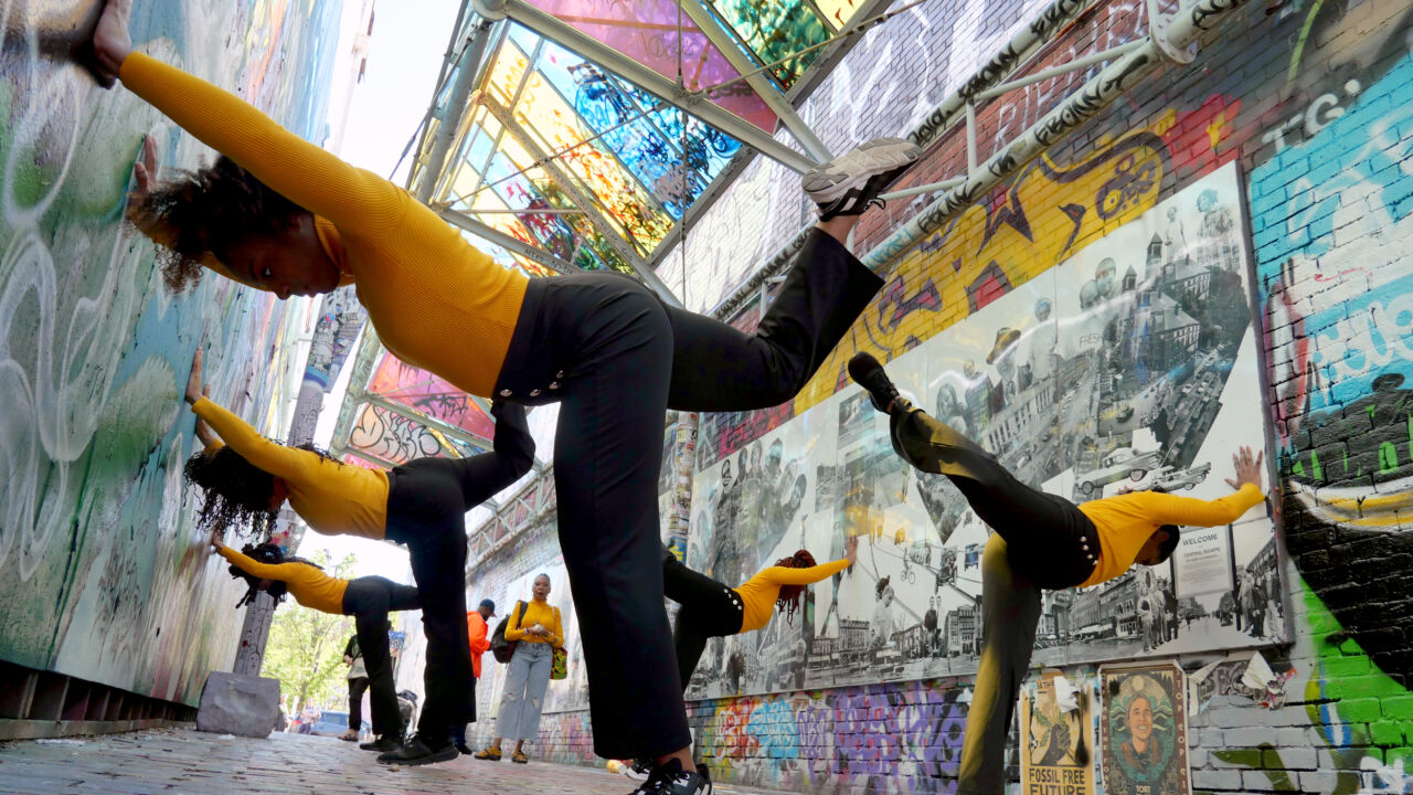 Four dancers with black pants and yellow turtlenecks, holding a pose - bent forward with arms extended and one foot raised up behind them, in a colorfully painted, bright city alleyway