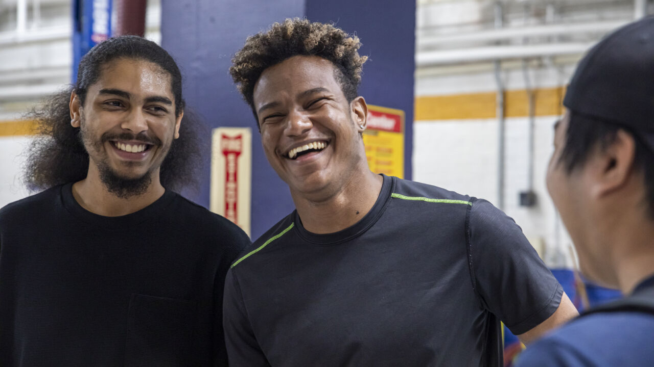 Two young men with big smiles facing the viewer, and a third young man in profile looking at them, and also appearing to smile