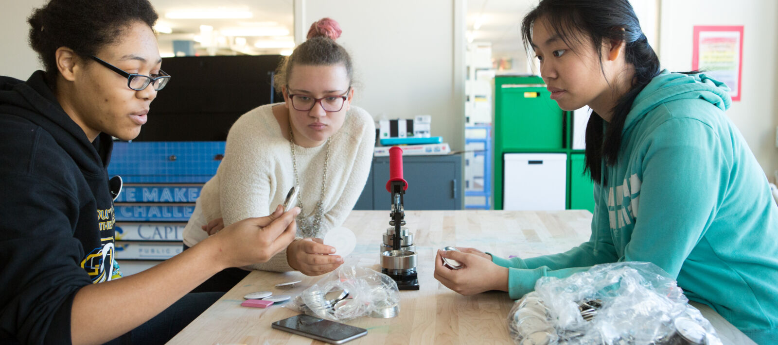 Three girls makes buttons in a high school classroom.