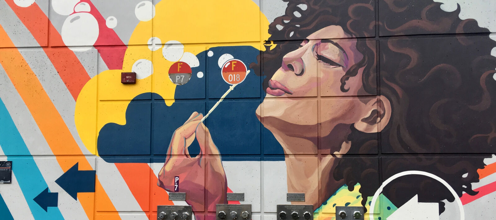 Colorful mural of woman with brown hair and red lipstick blowing white bubbles across a rainbow.