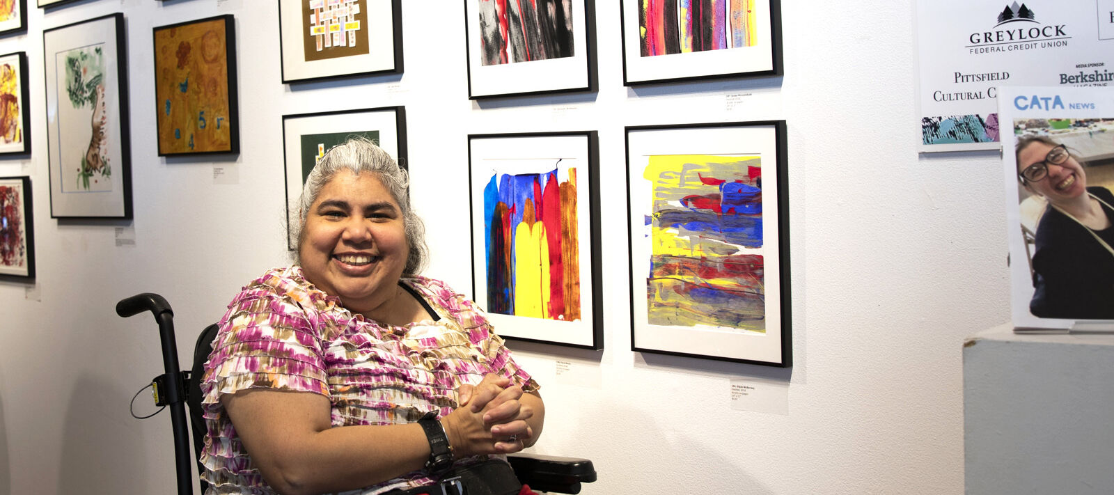 Woman sitting in a wheelchair in front of a wall of artwork, smiling
