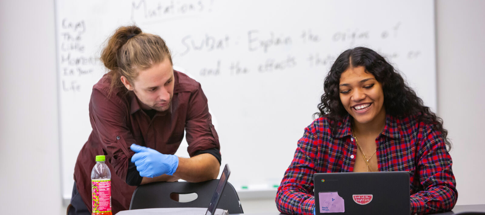 A student smiling at their computer while they work and a person beside them as they work.
