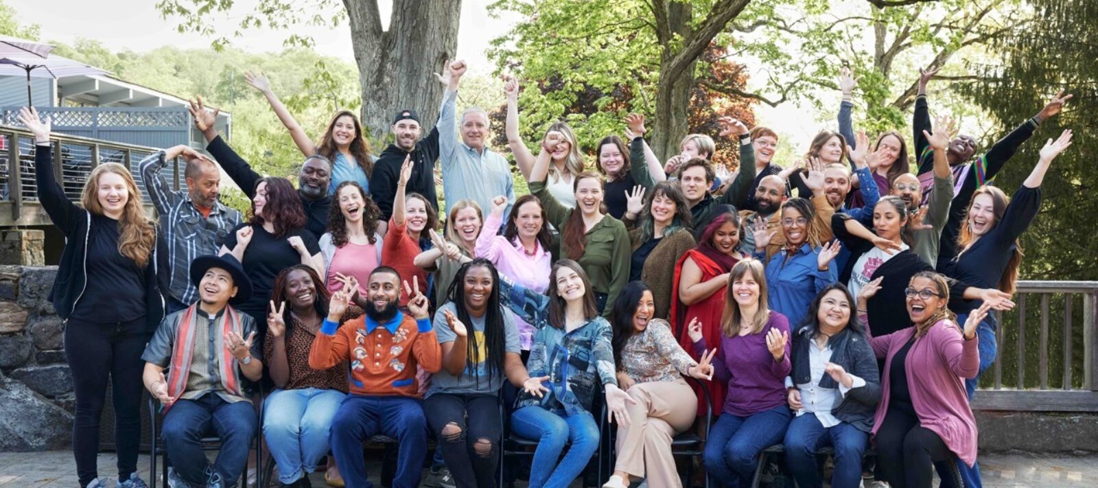 A diverse group of forty smiling people, many with hands raised, on a stone patio in front of green trees