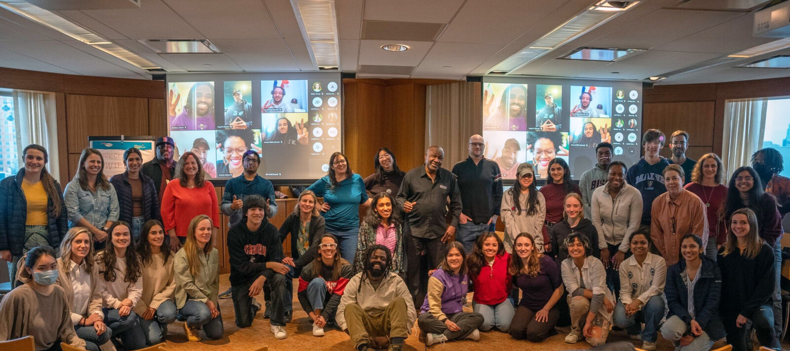 A diverse group of approximately 30 people smiling and posing in a conference room, standing in front of two projection screens with several additional people joining virtually.