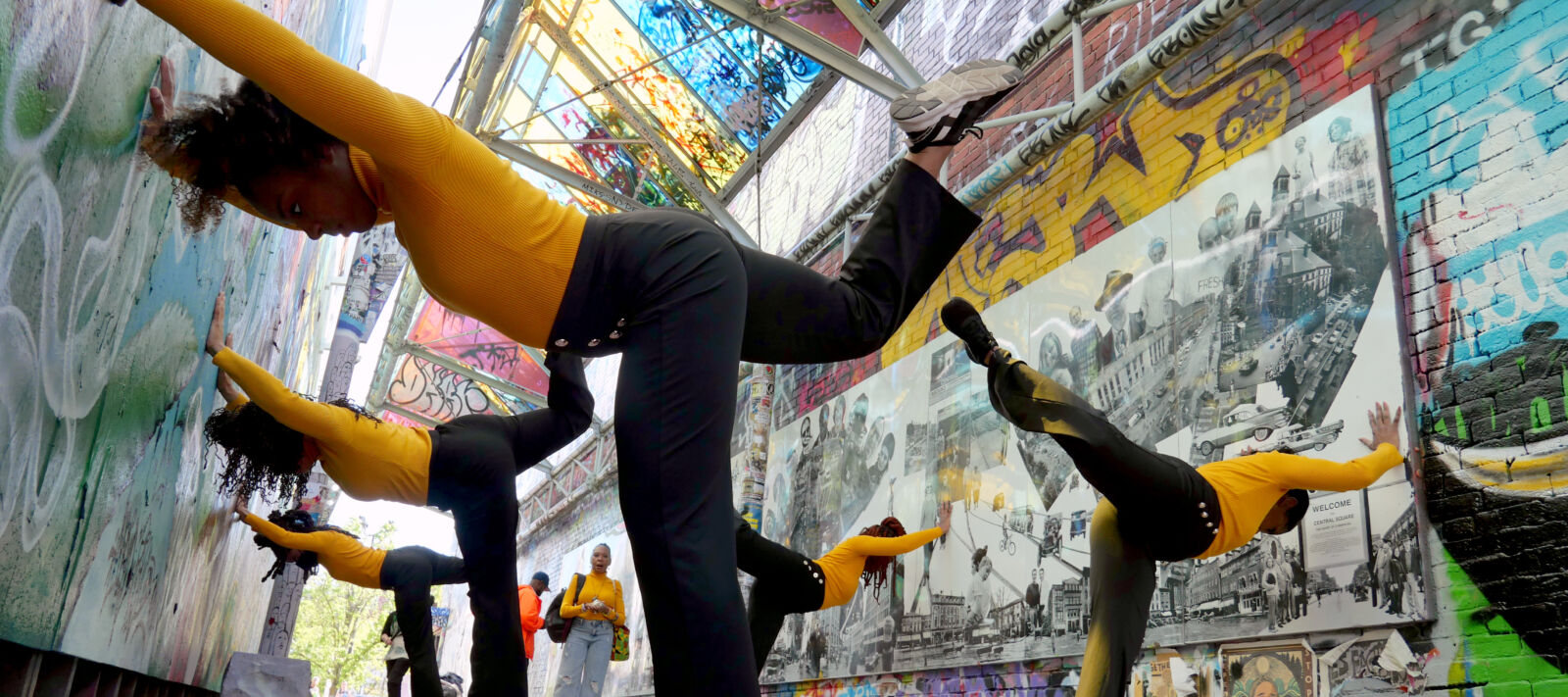 Four dancers with black pants and yellow turtlenecks, holding a pose - bent forward with arms extended and one foot raised up behind them, in a colorfully painted, bright city alleyway