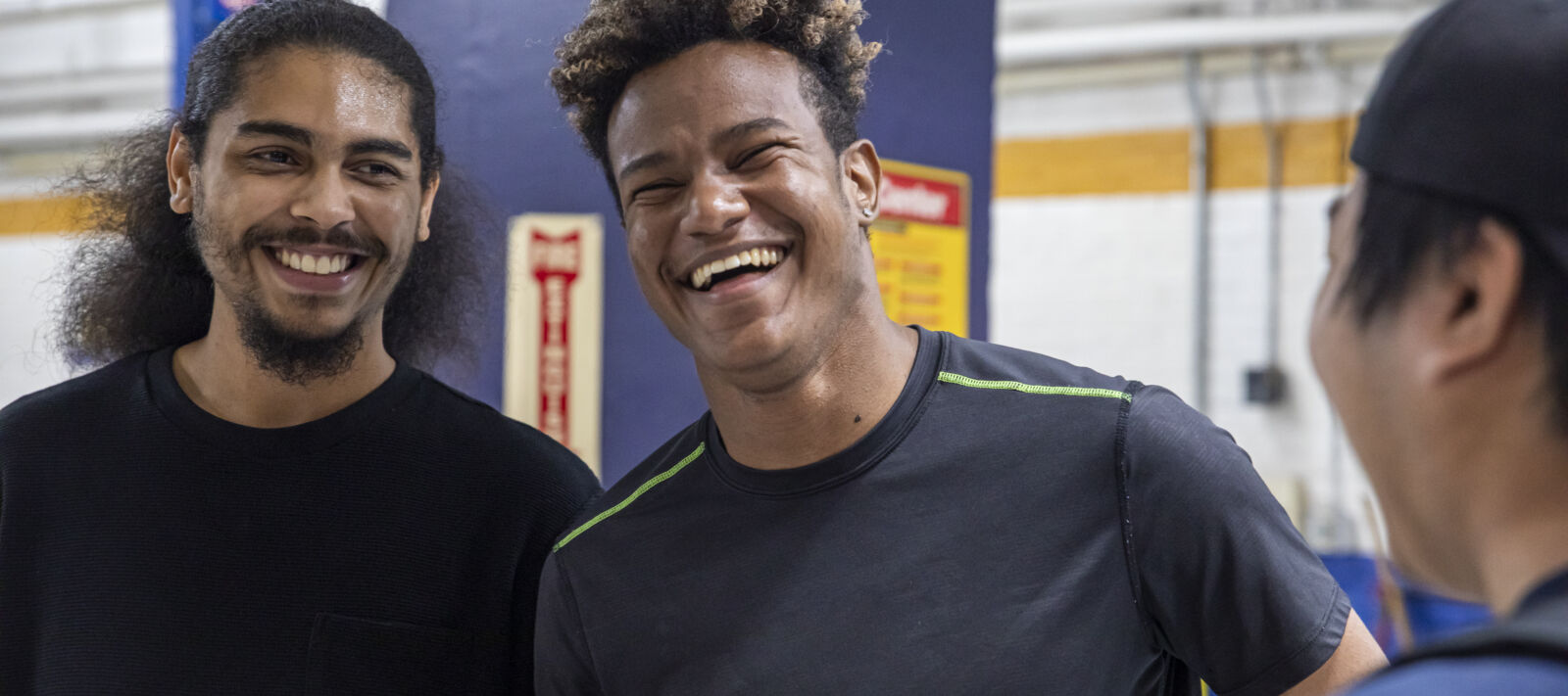 Two young men with big smiles facing the viewer, and a third young man in profile looking at them, and also appearing to smile