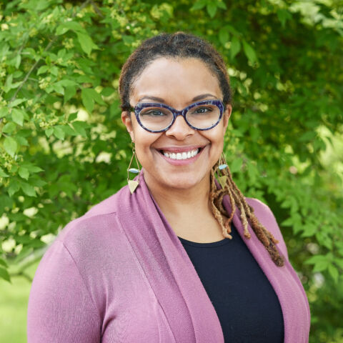 A profile photo of a smiling woman with black-rimmed glasses in front of a leafy green background