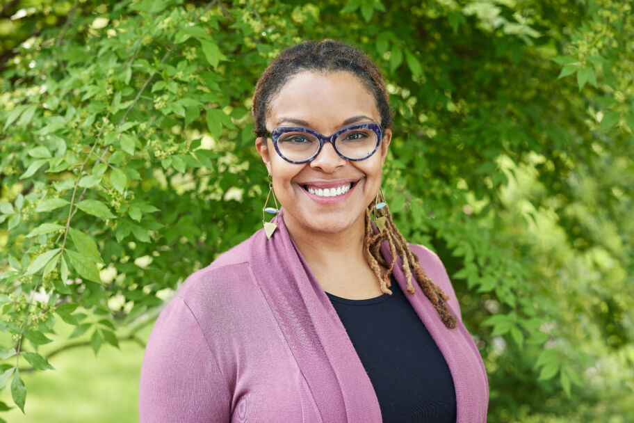 A profile photo of a smiling woman with black-rimmed glasses in front of a leafy green background
