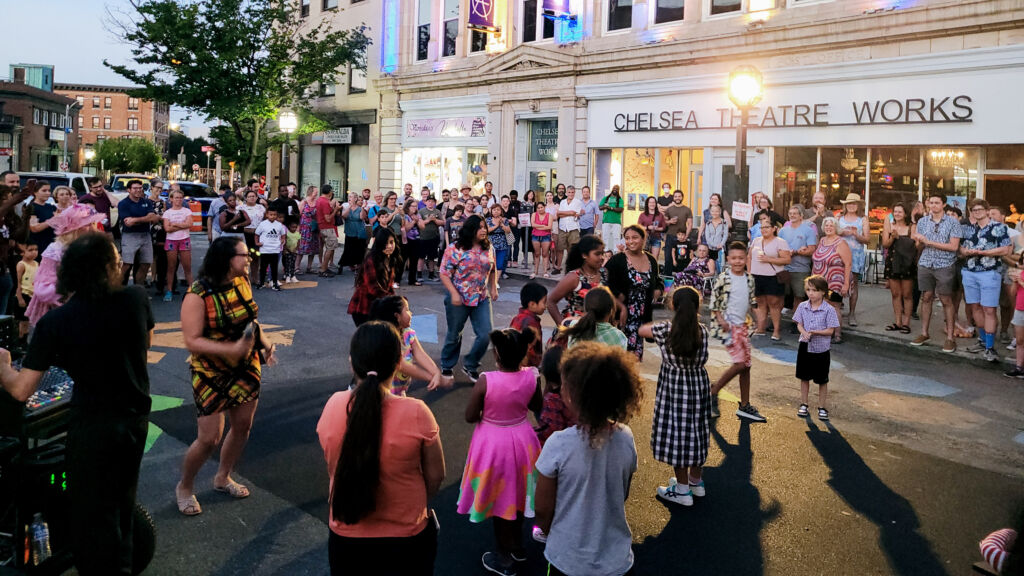 In the early evening, a group of people, including children are dancing in a street that is closed to cars and in front of the Chelsea Theater in Chelsea, MA.