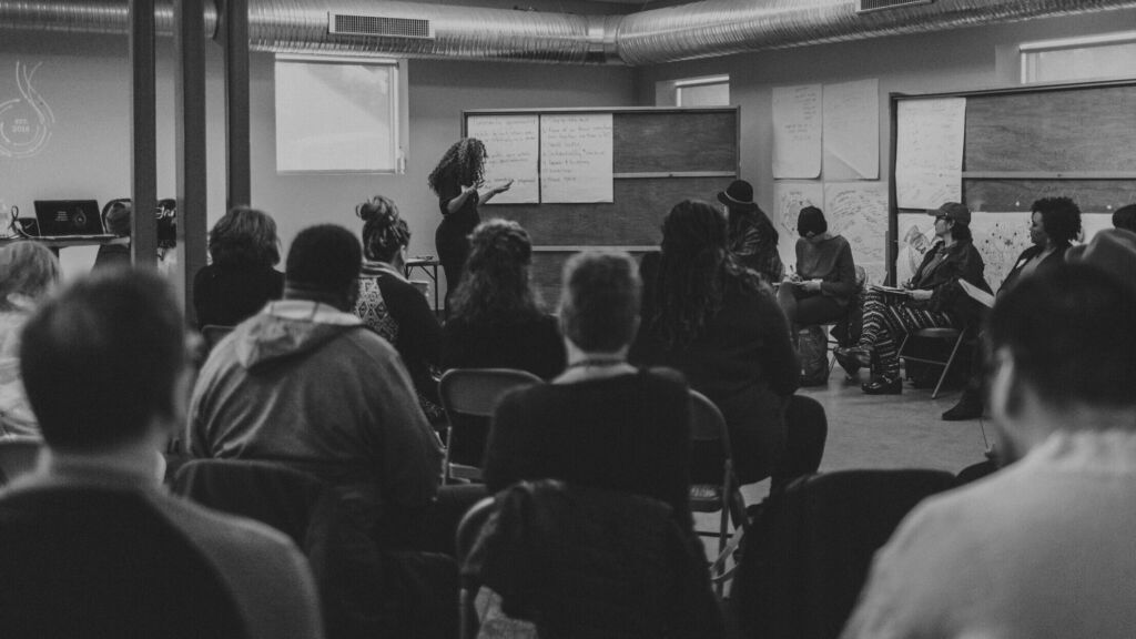 A black and white photo of a group of people sitting in a room watching a woman present and point to a flip chart