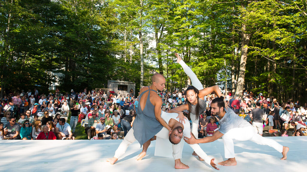 Four barefoot dancers wearing white pants and gray tops perform on stage while a large audience sitting outside in a serene, wooded setting watches.