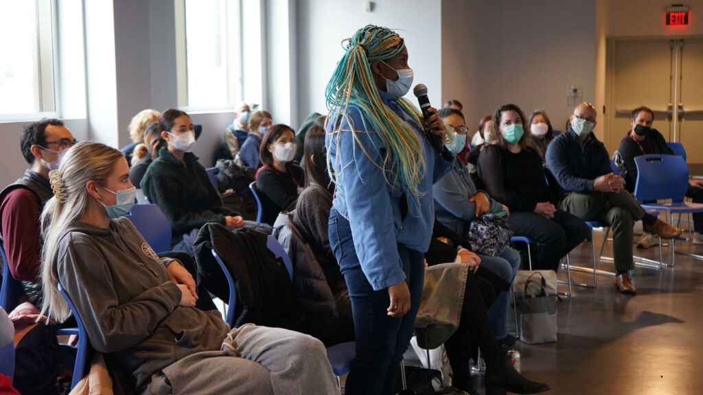 A woman with long braids wearing a medical mask and a sweatshirt stands up from within a seated crowd and speaks into a microphone.