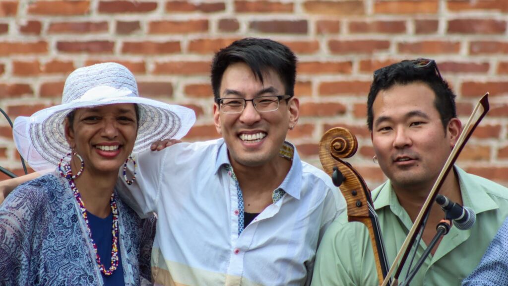 Photo showing one woman and two men standing outside in front of a brick wall. They are all smiling. The woman is wearing a white sun hat and one man is holding a violin.