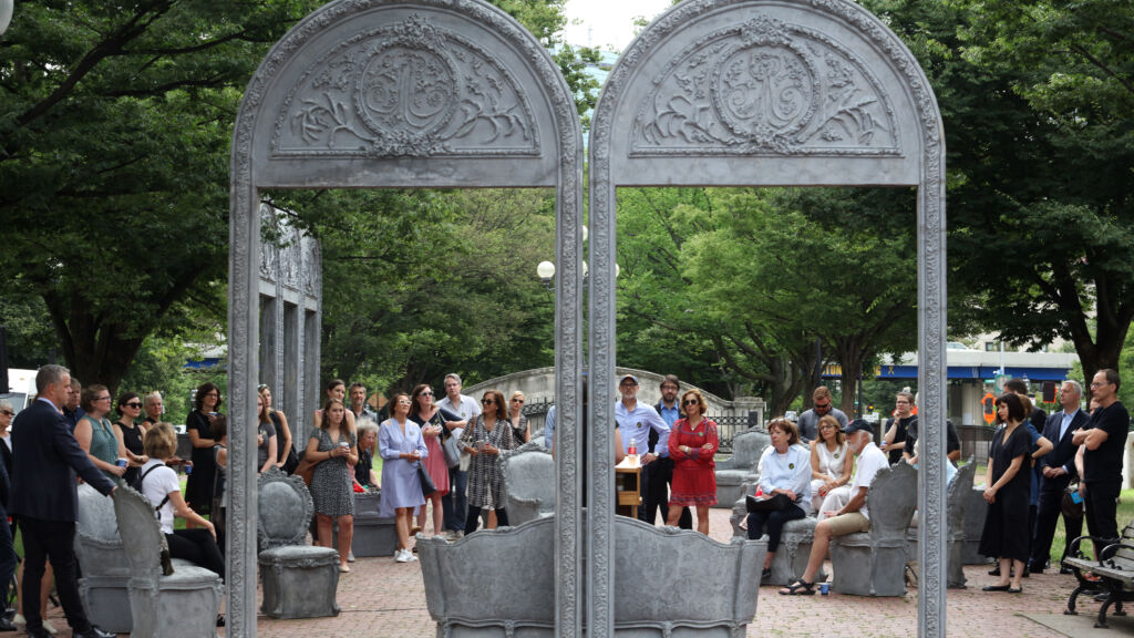 A group of people are standing in a park are looking at a public art exhibit by American visual artist, Liz Glynn.
