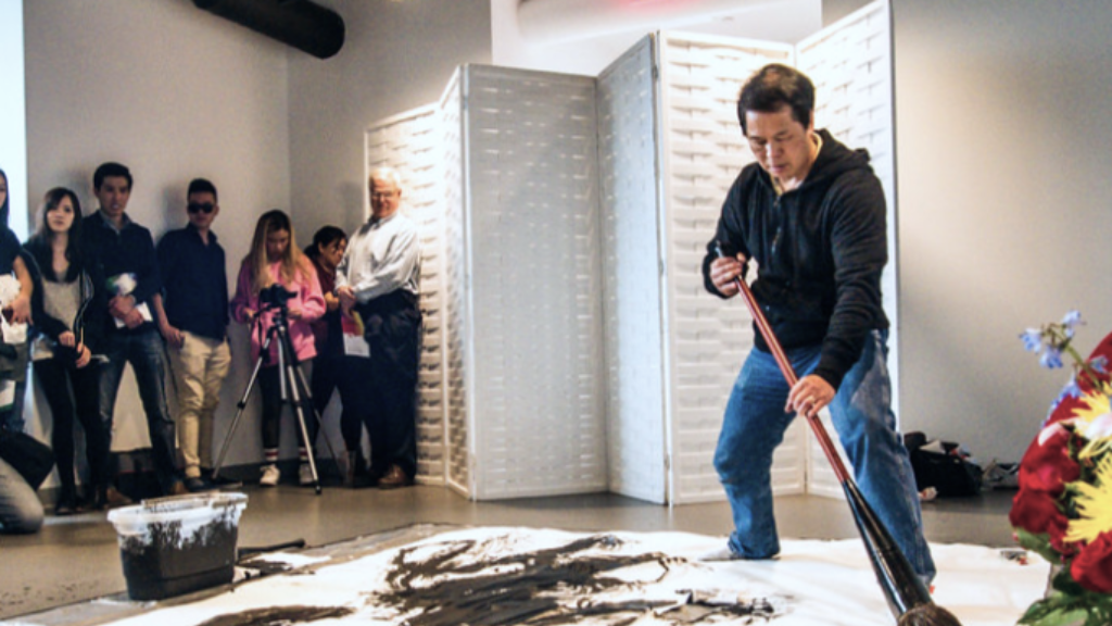 A Chinese man wearing a black sweatshirt paints with a large brush black paint across a white canvas on the floor. A crowd looks on including one woman with a videocamera.