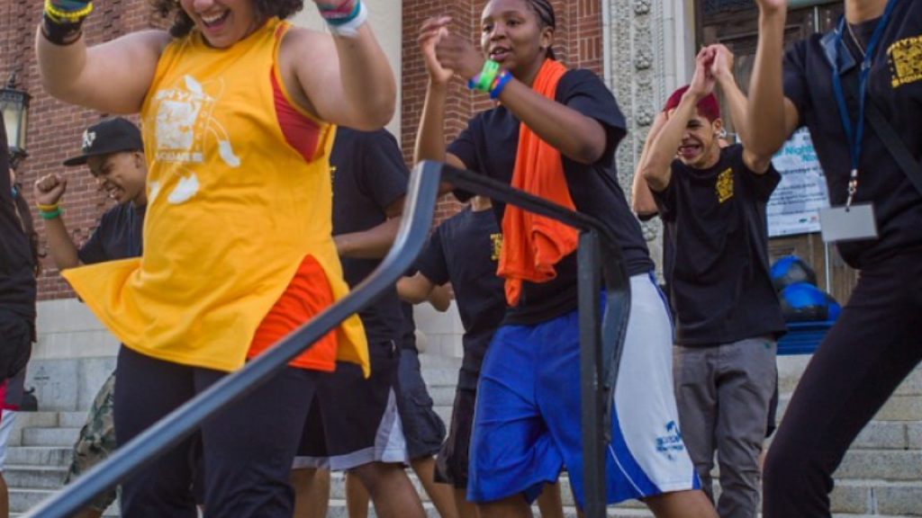 A group of teenagers happily perform a dance outside a brick building on a set of concrete steps.