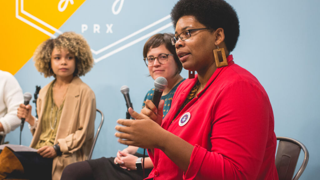 Three women are sitting inside the Podcast Garage and speaking on a panel. One Black woman in the foreground wears a red blazer speaks into a handheld microphone.