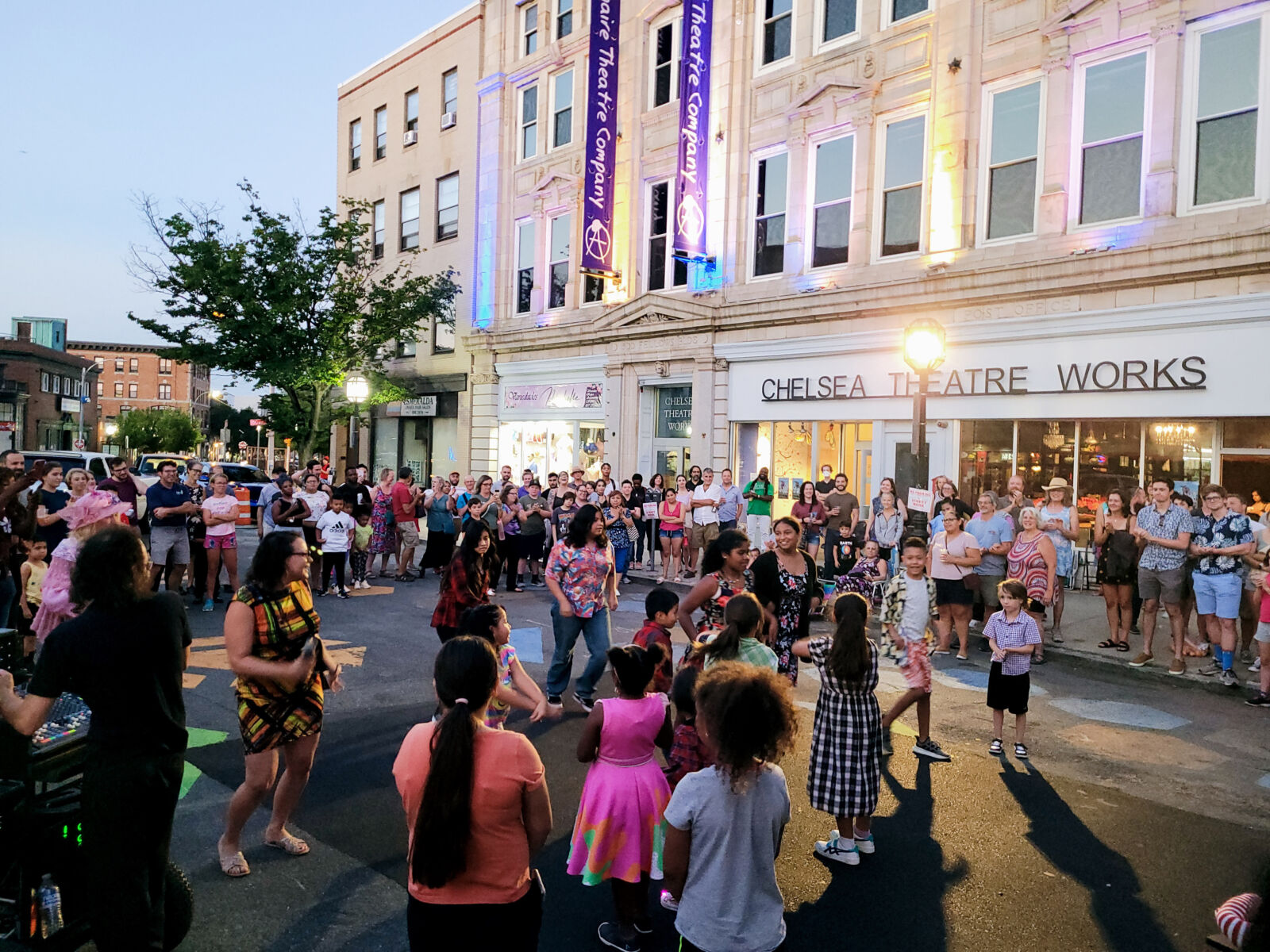In the early evening, a group of people, including children are dancing in a street that is closed to cars and in front of the Chelsea Theater in Chelsea, MA.