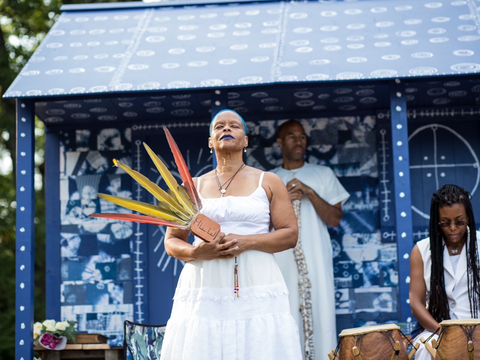 A Black woman with short blue hair and blue lipstick stands in front of a small blue house with people behind her singing and playing drums. The woman is closing her eyes and looking up to the sky.