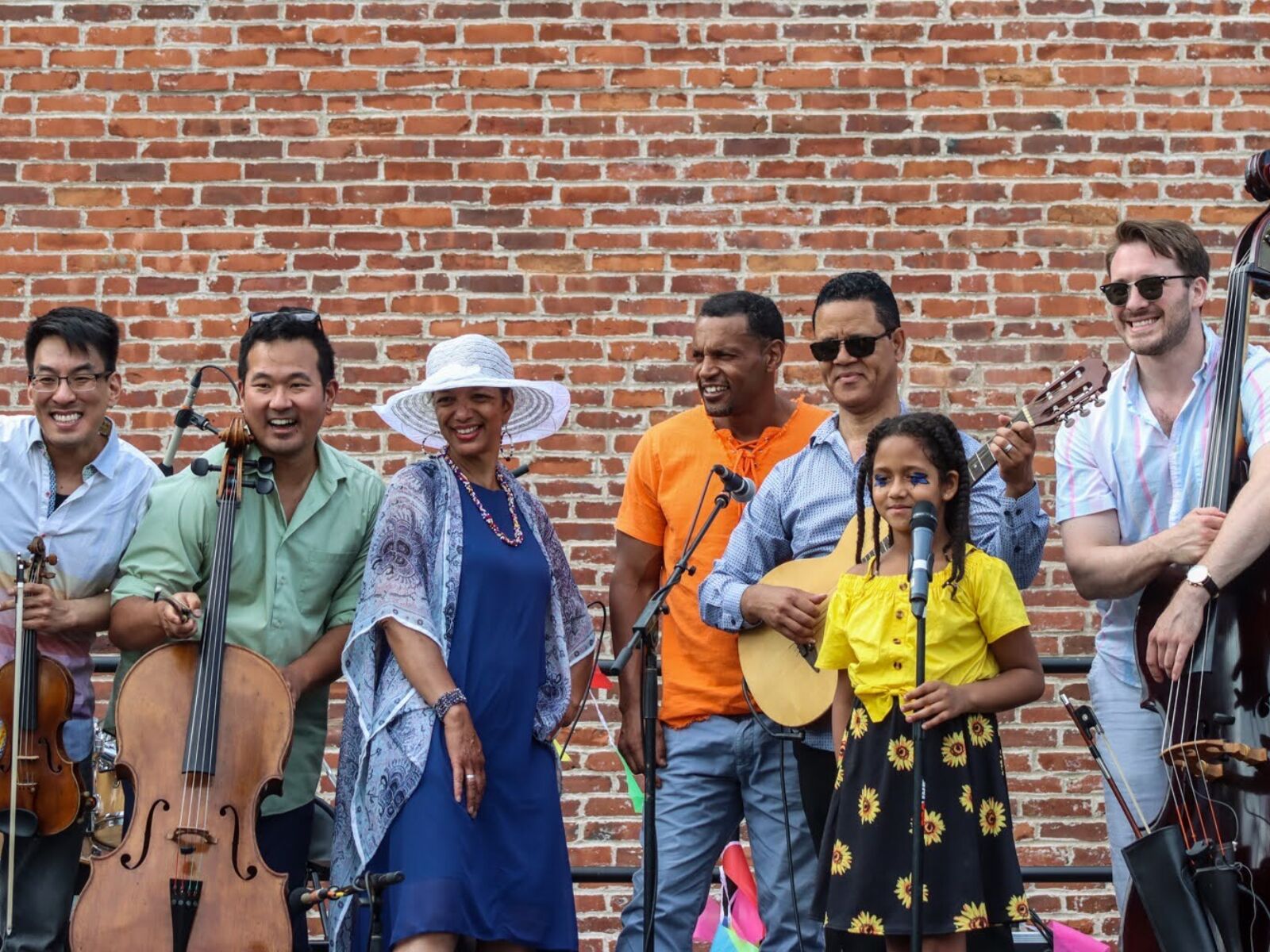 A diverse group of smiling musicians pose in front of a brick wall, some are holding string instruments and others are standing behind microphones.