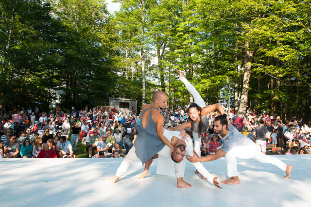 Four barefoot dancers wearing white pants and gray tops perform on stage while a large audience sitting outside in a serene, wooded setting watches.