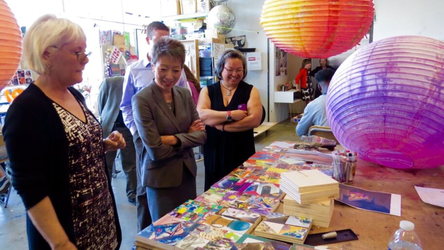 A large group of artists sit together in a room. The space is warm and bright, and seems full of energy. Those seated on the floor, on pillows, and in chairs look excitedly at a central presenter, who faces the group and gestures openly like a conductor..
