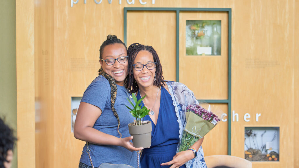 Two Black women wearing glasses and holding flowers are embracing and smiling