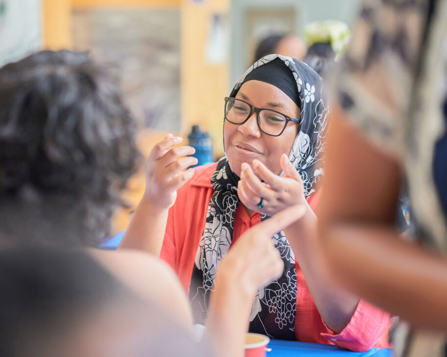 Candid photo of a Black woman wearing a headscarf and glasses sitting at a table and talking to other people.
