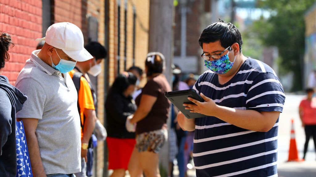 A man in a mask holds an iPad as he gathers information from a community member as part of a Census 2020 outreach effort. Others appear in the background and are dressed casually for summer weather, standing in small groups outside.