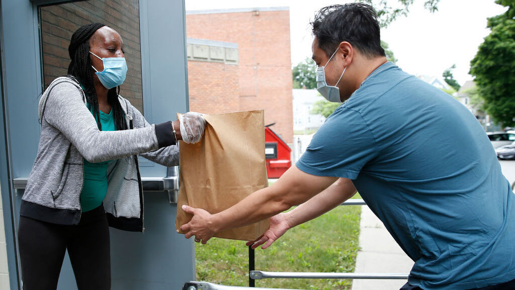 A woman wearing a face mask and gloves hands a paper bag full of food to a man wearing a face mask. They stand at a side exit of a school.